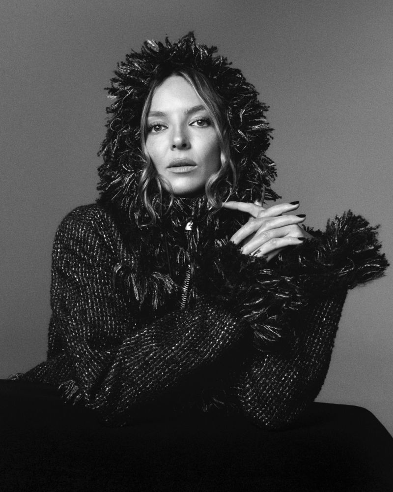Jodie Comer wears a heavy tweed jacket with a textured fringed hood and cuffs, clasping her hands against a grey studio backdrop.