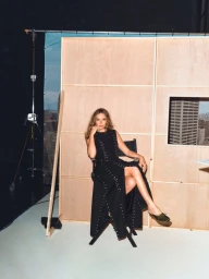 Jodie Comer seated in a black dress in a studio set with wooden flat panels and studio equipment.
