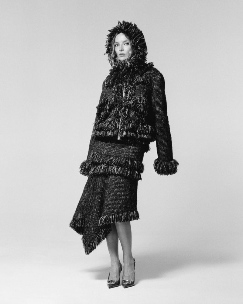 Jodie Comer standing in a dark textured fringe hooded jacket and matching asymmetric skirt on a seamless studio backdrop.