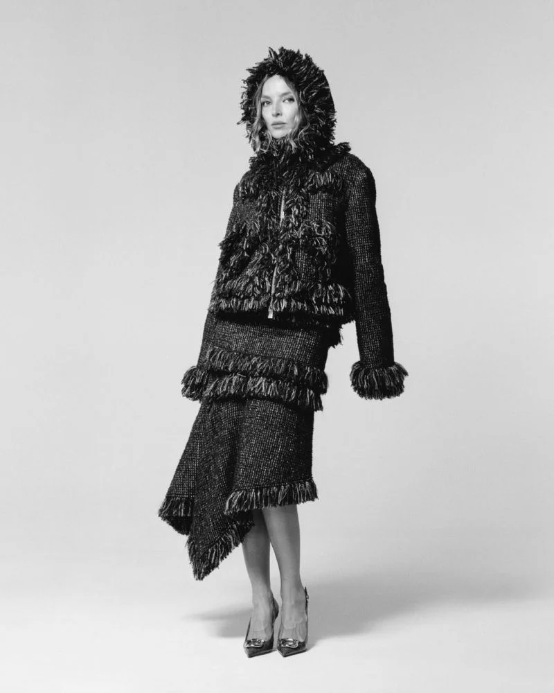 Jodie Comer standing in a dark textured fringe hooded jacket and matching asymmetric skirt on a seamless studio backdrop.