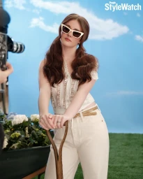 Harlow Jane wearing white cat-eye sunglasses and cream trousers, posing with garden tools against a painted sky studio backdrop.
