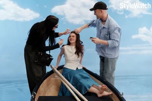Harlow Jane seated barefoot in a wooden prop rowboat as crew members adjust her hair and makeup on a studio set.