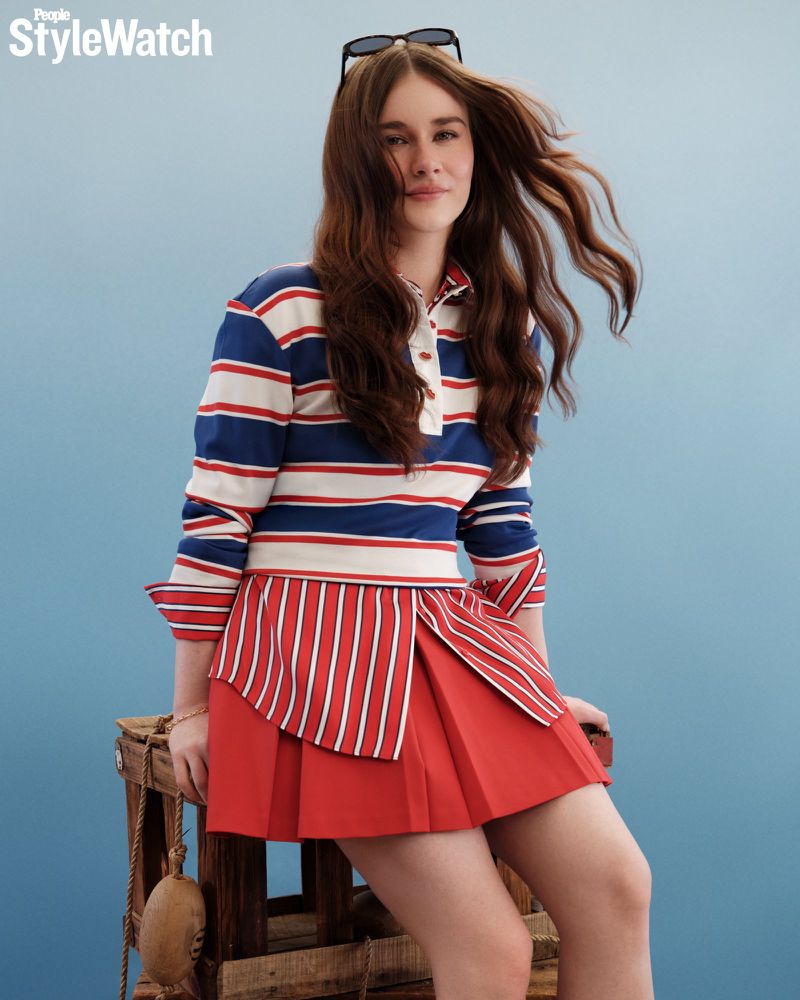 Harlow Jane seated on a wooden crate wearing a red and blue striped long-sleeve polo and red pleated skirt against a blue studio backdrop.