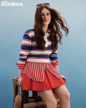 Harlow Jane seated on a wooden crate wearing a red and blue striped long-sleeve polo and red pleated skirt against a blue studio backdrop.