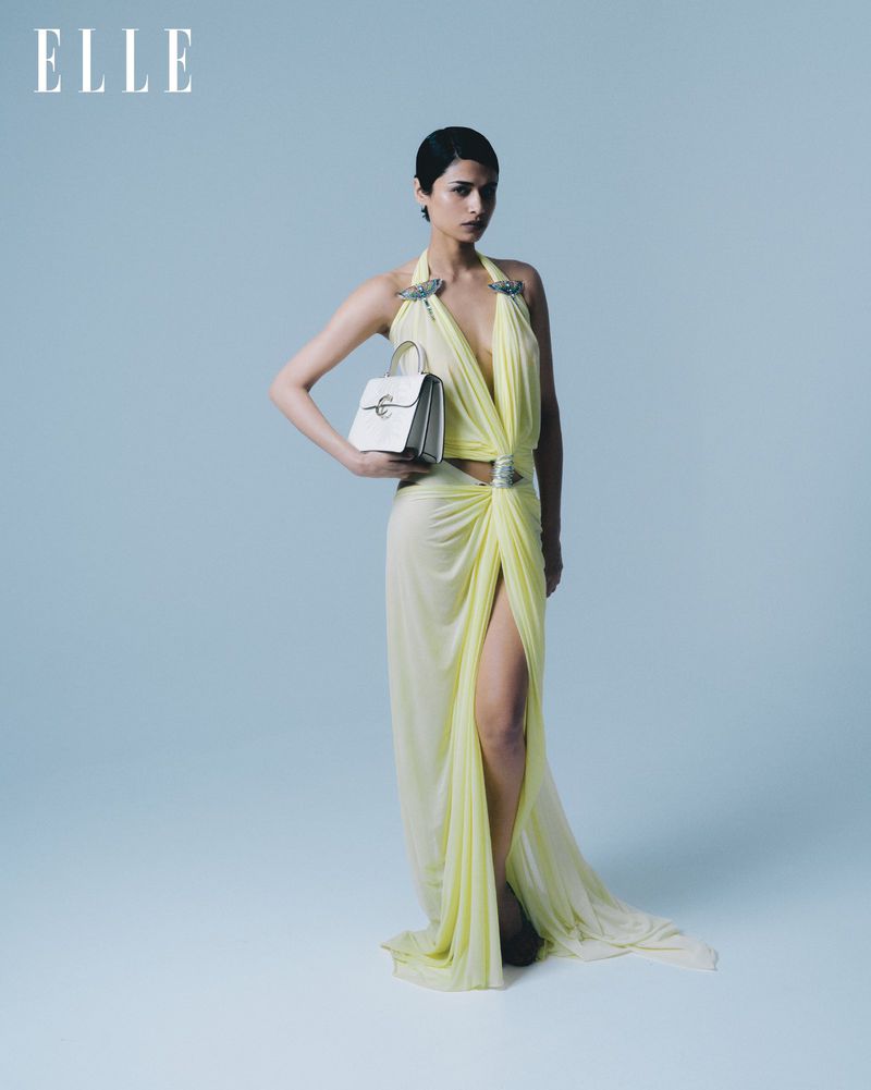 Model Kirandeep Chahal posing in a pale yellow draped halter gown with a high slit while holding a white structured handbag.