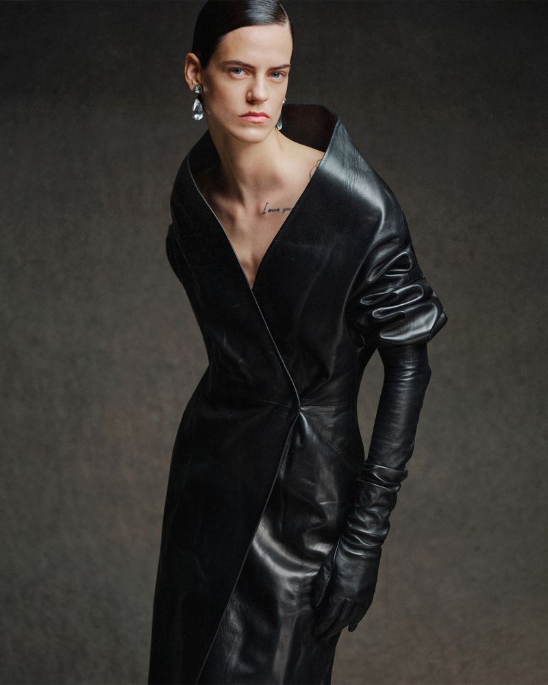Model Miriam Sanchez wearing a structured black leather coat dress with long leather gloves against a dark textured studio backdrop.