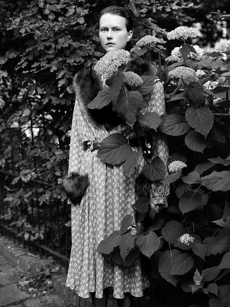 Julie Hoomans posing in a fur-trimmed patterned dress beside hydrangea bushes in a black and white editorial.