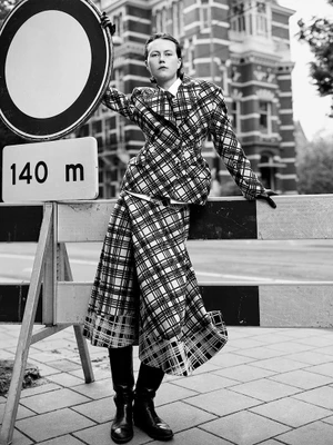 Julie Hoomans in a plaid suit and black boots leans on a road barricade and circular sign on a city street.