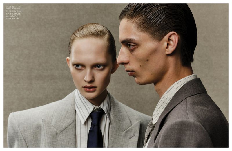 Fashion editorial shot of a young adult couple in grey suits and striped shirts in a photo studio with neutral background, gazing sideways.
