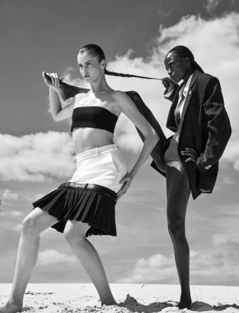 Two fashion models pose sculpturally in monochrome on sand under a cloudy sky, one holding the other's long braided hair.