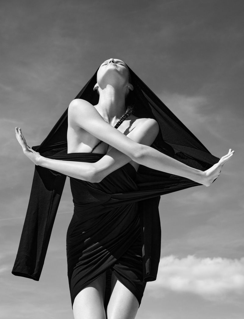 Low-angle black-and-white fashion portrait of model Lesia Havrylets in a draped black dress posing dynamically against the open sky.