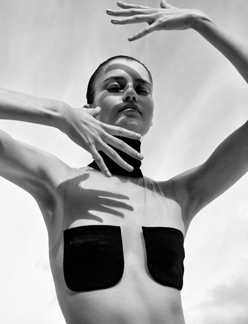Black and white low-angle portrait of a woman framing her face with raised hands that cast sharp shadows across her features.