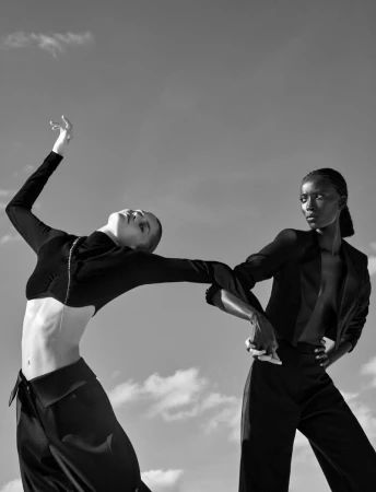 Two fashion models in black outfits posing dynamically holding hands outdoors under a bright sky in black and white.