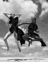 Two models in black sculptural swimwear leaping mid-air above white sand against a cloudy sky in black and white.