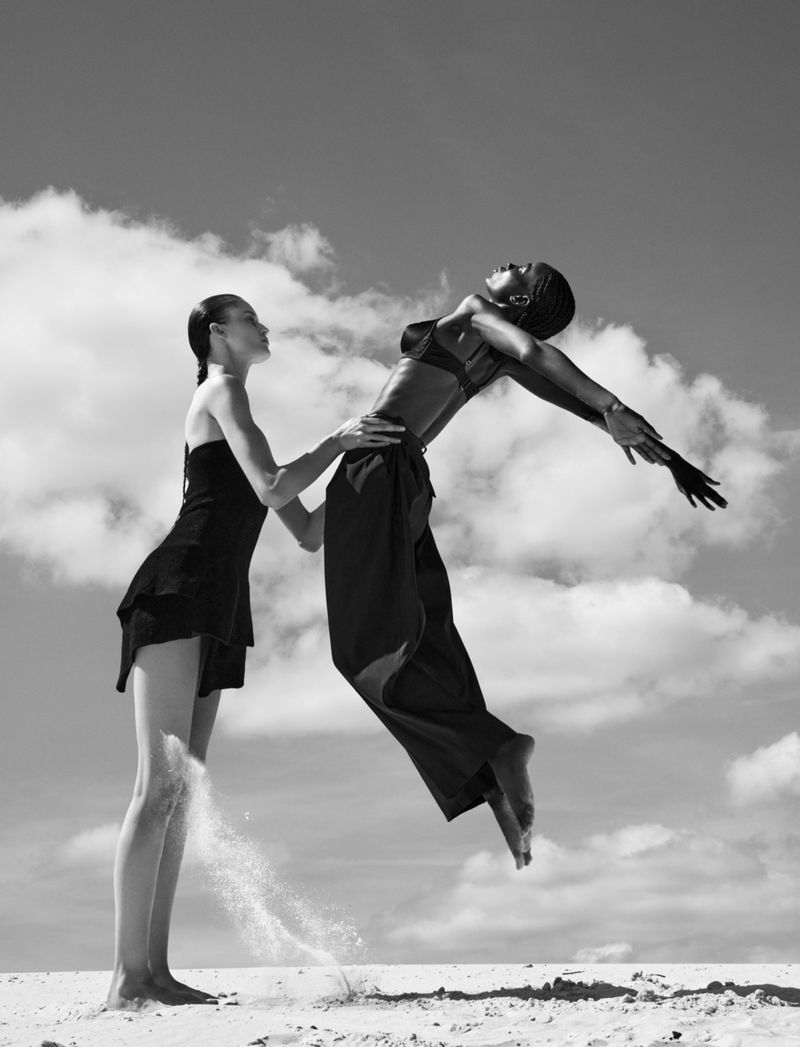 Black and white photo of two female models on sand, one supporting the other mid-air with arms arched backwards against a cloudy sky.