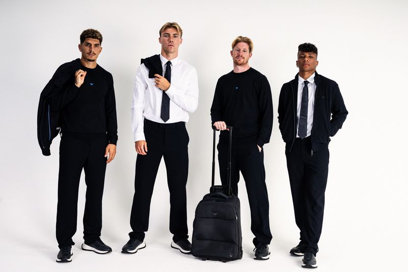 Four men in Emporio Armani black and white tailored menswear with a black rolling suitcase against a white seamless backdrop.