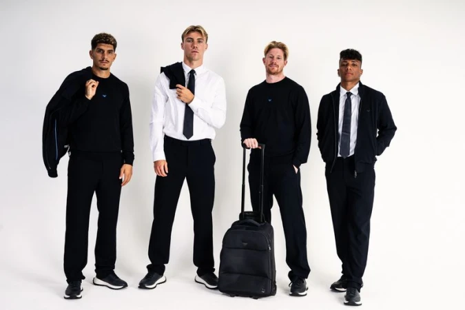 Four men in Emporio Armani black and white tailored menswear with a black rolling suitcase against a white seamless backdrop.