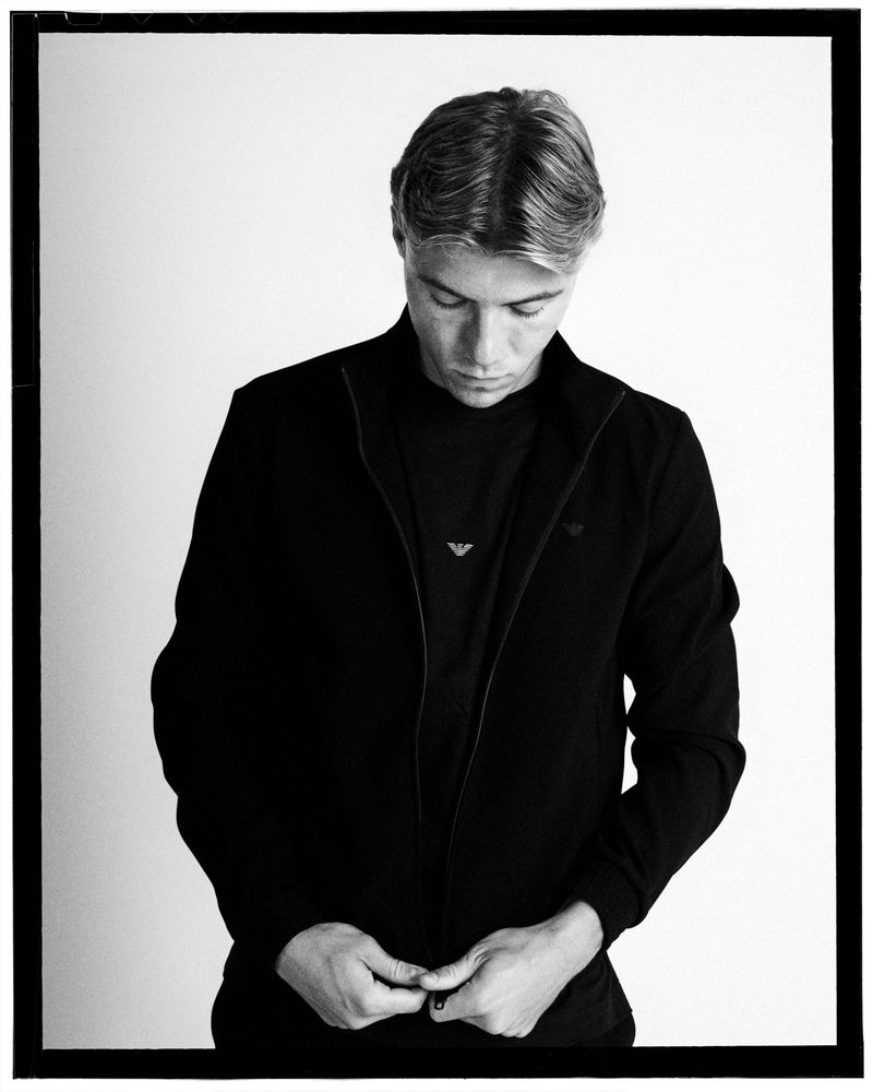 Monochrome portrait of a young man in a black Emporio Armani zip jacket looking downward against a white studio background.