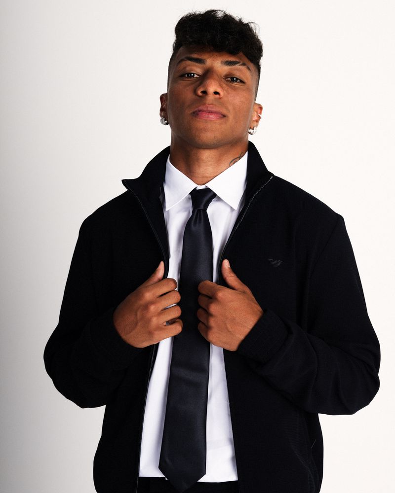Young man posing in a black zip-up jacket over a white dress shirt and dark necktie against a white backdrop.