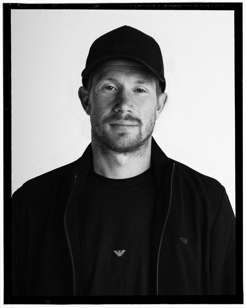 Black and white portrait of a man wearing a dark baseball cap, jacket, and Emporio Armani logo shirt against a white backdrop.