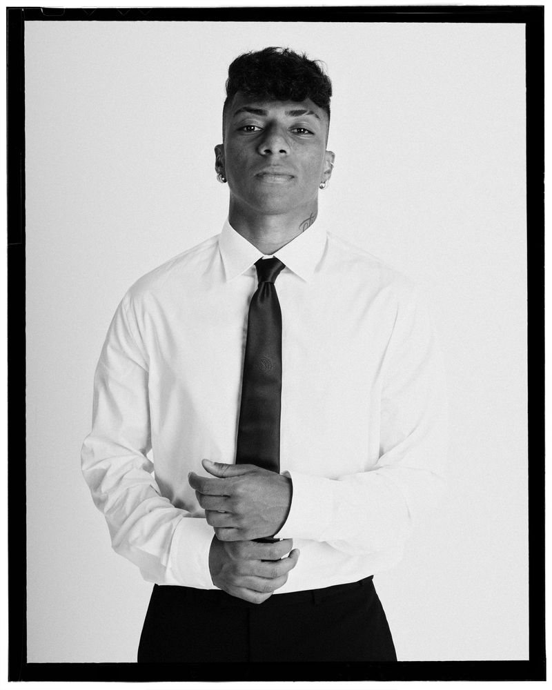 Man in a white dress shirt and dark tie adjusting his cuff against a plain white studio background.