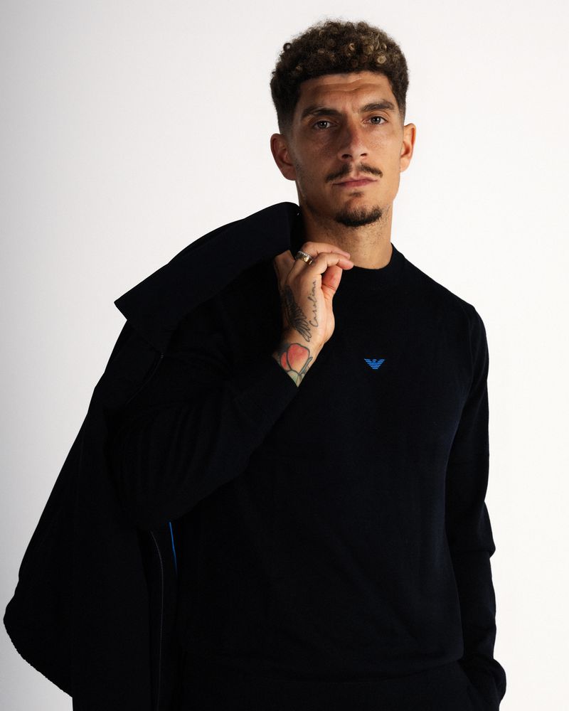 Giovanni Di Lorenzo in a black Emporio Armani sweater holding a blazer over his shoulder against a white studio background.