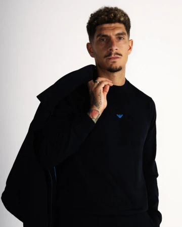 Giovanni Di Lorenzo in a black Emporio Armani sweater holding a blazer over his shoulder against a white studio background.
