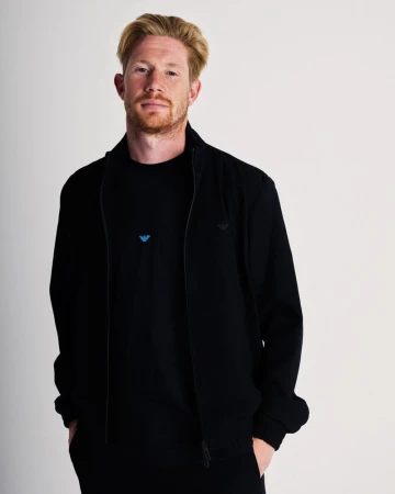 Man with strawberry blonde hair wearing a black Emporio Armani zip-up jacket and black shirt against a plain white background.