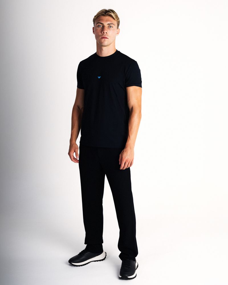 Blonde man wearing a black crewneck t-shirt, black trousers, and sneakers standing against a white studio background.