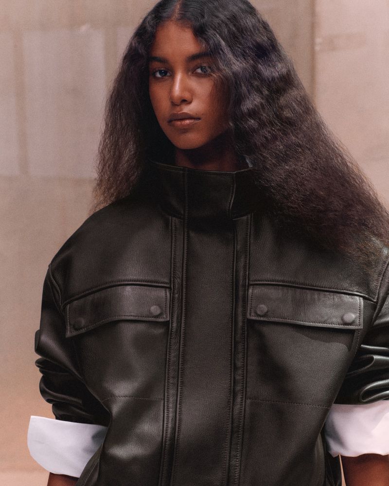 Model Luna Yohannan wearing an oversized black leather high-neck jacket against a warm neutral stone wall.