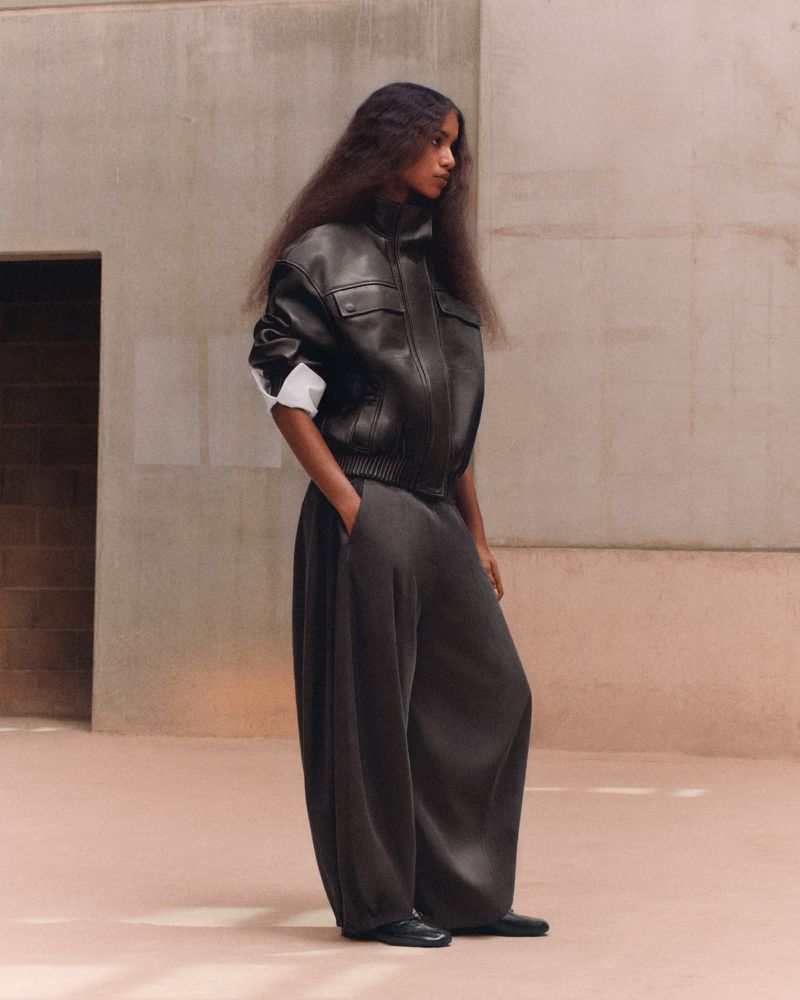 Model standing in profile wearing a black leather bomber jacket and oversized dark balloon trousers against concrete walls.