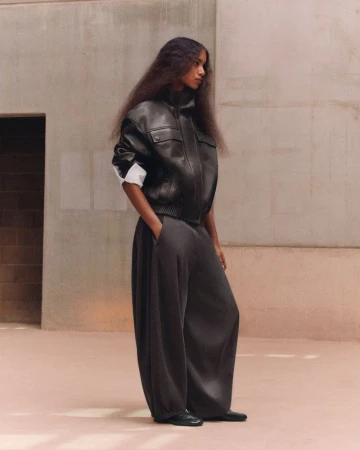 Model standing in profile wearing a black leather bomber jacket and oversized dark balloon trousers against concrete walls.