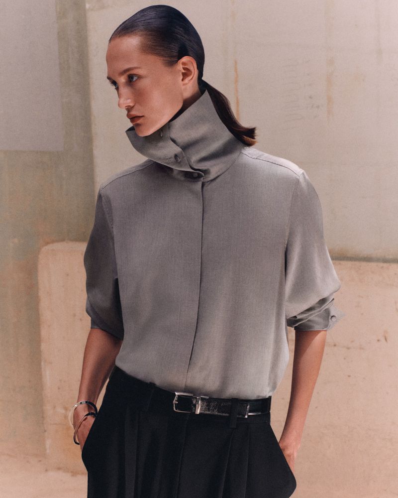 Model wearing a high collar grey buttoned shirt and black pleated trousers with a black belt against a neutral textured wall.