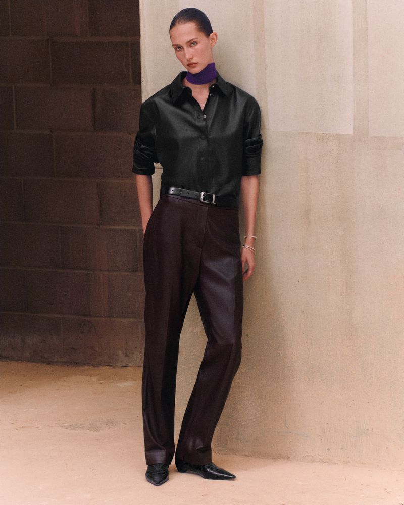 Model leaning against a stone wall wearing a black long-sleeve shirt, purple neckband, and dark tailored trousers.