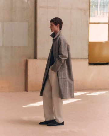 Model standing in profile wearing a long plaid coat, cream trousers, and dark shoes in a sunlit industrial space.