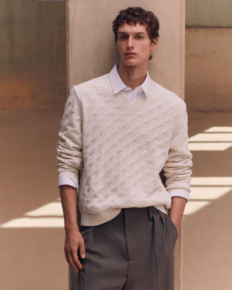 A man wearing a cream textured V-neck sweater over a white collared shirt and charcoal pleated trousers leaning against a column.