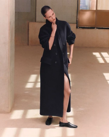 Model posing in a long black double-breasted maxi coat and black flat shoes against warm minimalist architectural walls with sunlit shadow patterns.