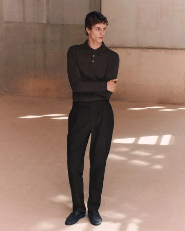 Male model wearing a dark long-sleeve knit polo shirt and black tailored trousers standing on a warm concrete floor.