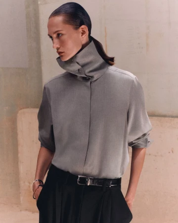 Model wearing a grey high-neck buttoned shirt tucked into black trousers with a belt against a textured neutral background.