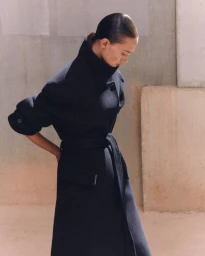 Woman wearing a structured dark navy belted coat with a high collar, posed against a warm neutral textured studio wall.