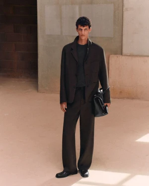 Model standing in a relaxed dark tailored suit and black shoes while holding a black leather tote bag in a minimalist setting.