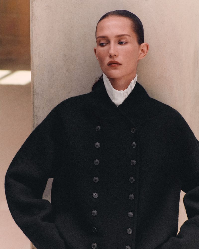 Model wearing a black double-breasted coat with a pleated white high collar posed against a neutral wall.