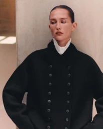 Model wearing a black double-breasted coat with a pleated white high collar posed against a neutral wall.