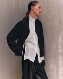 A woman in a black jacket and layered white high-collar tunic leans against a textured grey concrete wall.
