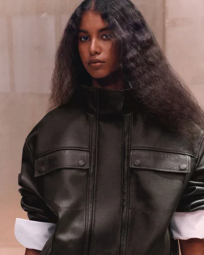 Model Luna Yohannan wearing an oversized black leather high-neck jacket with white cuffs against a warm neutral backdrop.