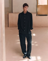 Male model wearing an oversized black button-up shirt and relaxed black trousers in a beige architectural interior.
