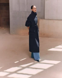 Model in a high-collar belted navy overcoat and wide-leg denim jeans standing in a sunlit architectural courtyard.