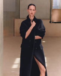 Model Vika Evseeva wearing a tailored long black double-breasted coat with a front slit in a warm minimalist interior setting.