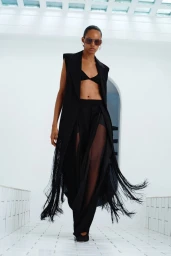 Model Sabryna Oliveira in a sleeveless black fringed coat, black bralette, and sunglasses in a white tiled interior.