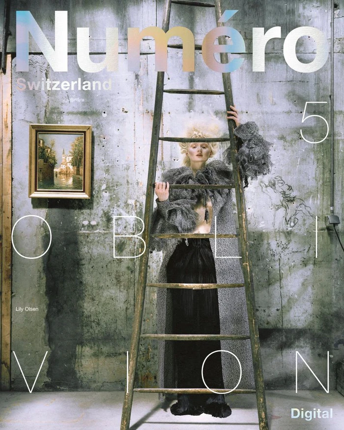 Fashion model in a voluminous grey ruffled cape standing behind a tall wooden ladder against a weathered concrete wall.